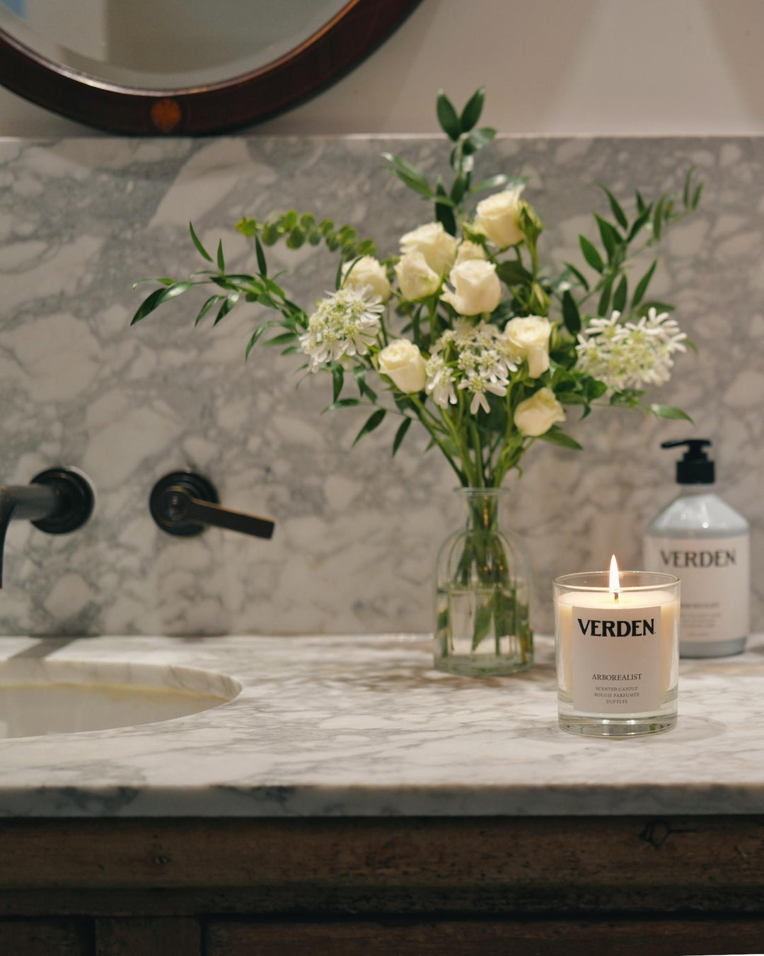 Marble bathroom basin with black wall-mounted taps, white roses in a glass vase and a lit Verden scented candle, styled for a cosy evening at home.