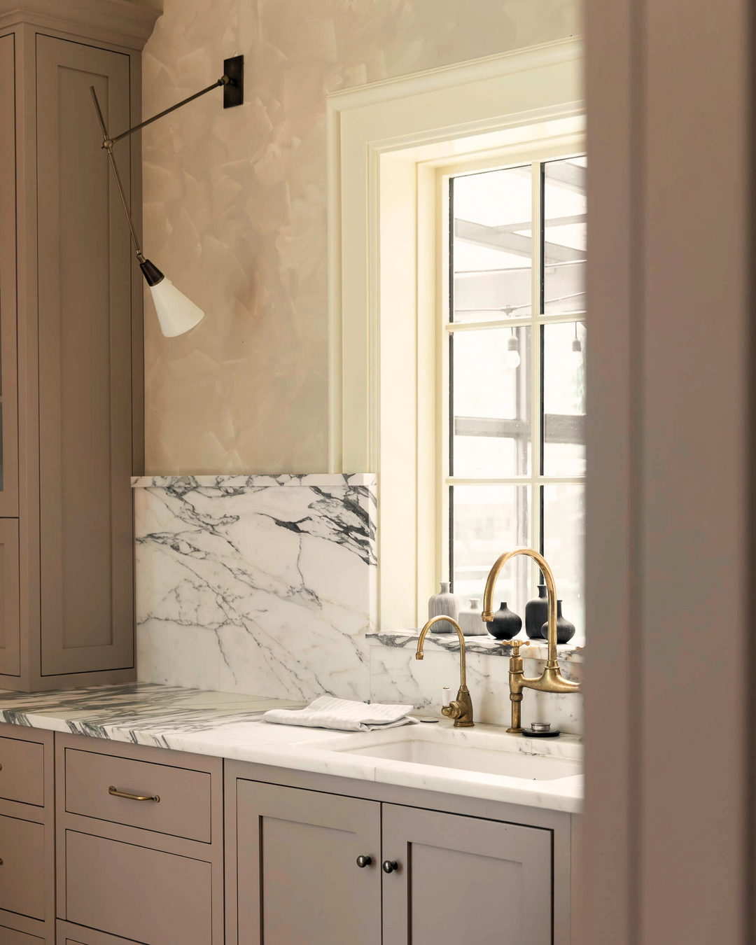 Pink kitchen with a marble splashback, brass tap and Shaker-style cabinets in warm taupe, a swing-arm wall light and a tall window letting in soft daylight.