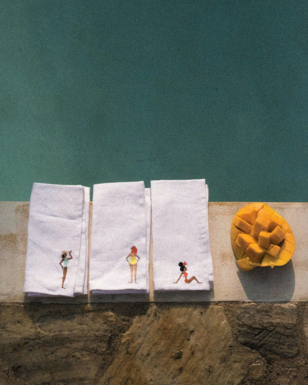 Three white linen napkins with embroidered designs of women in swimsuits on a stone ledge with a teal wall in the background.