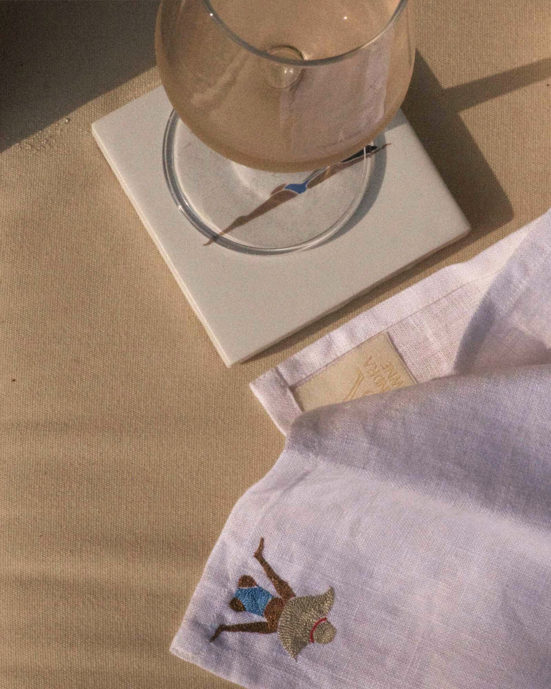 Glass on a white coaster with a napkin featuring an embroidered design on a beige surface.