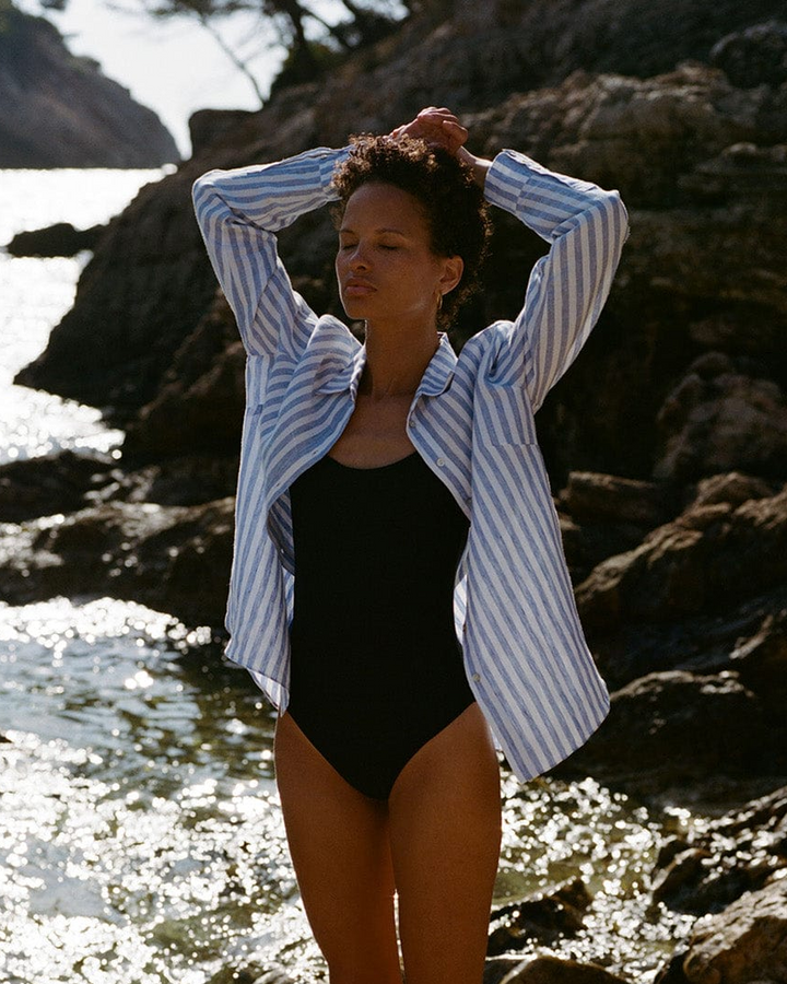 Woman in a black swimsuit and striped shirt standing by a rocky shoreline
