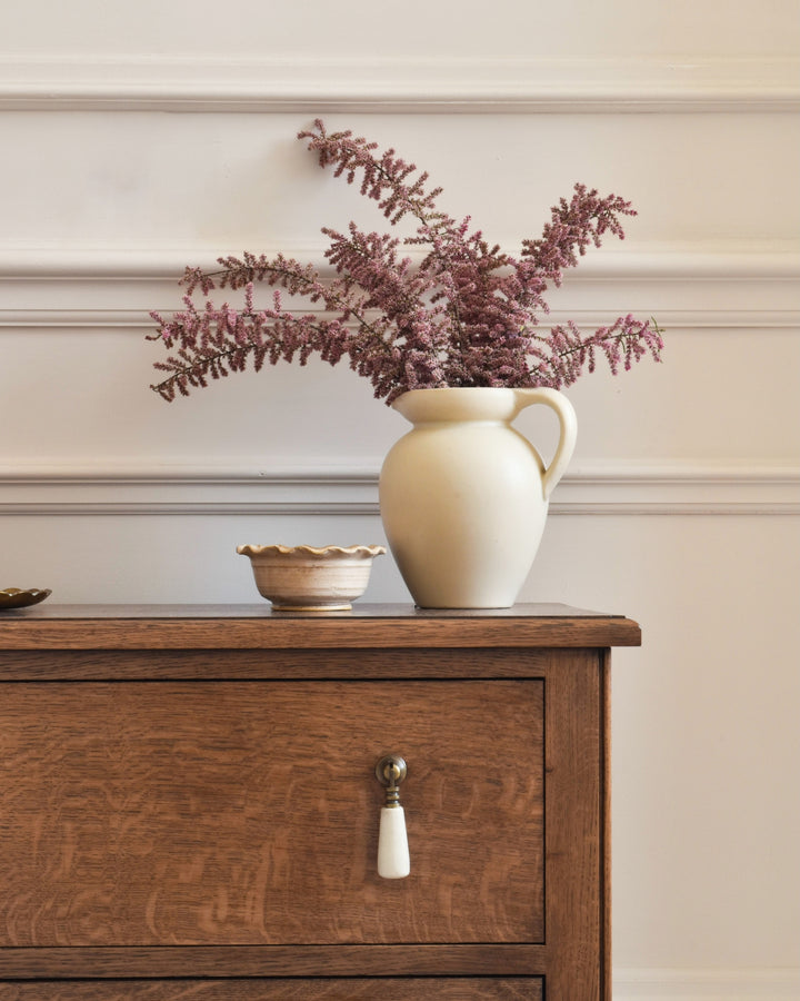 White pitcher with purple flowers on a wooden bedside table against a beige wall.