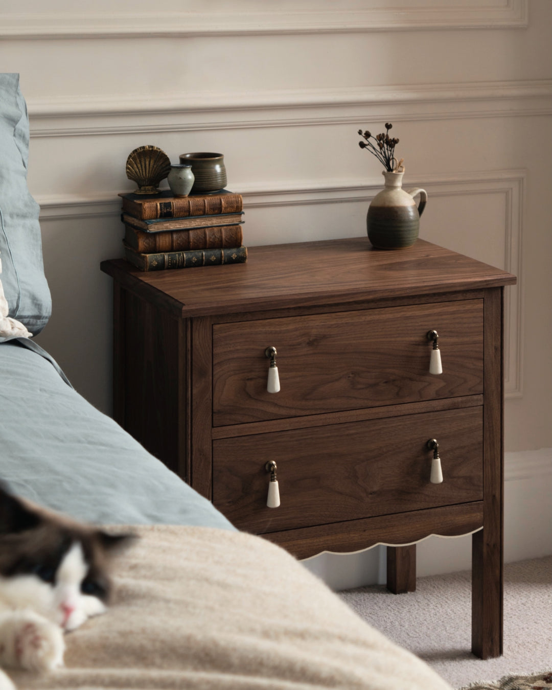 Wooden nightstand with two drawers, books, and a vase in a bedroom setting.