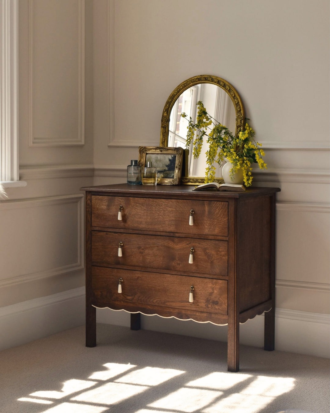 Wooden dresser with mirror and decorative items in a room with sunlight streaming through a window.