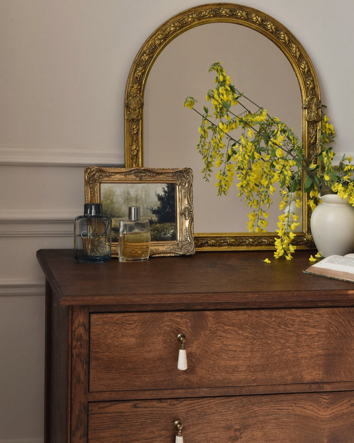 Wooden dresser with gold-framed mirror, yellow flowers, and decorative items.
