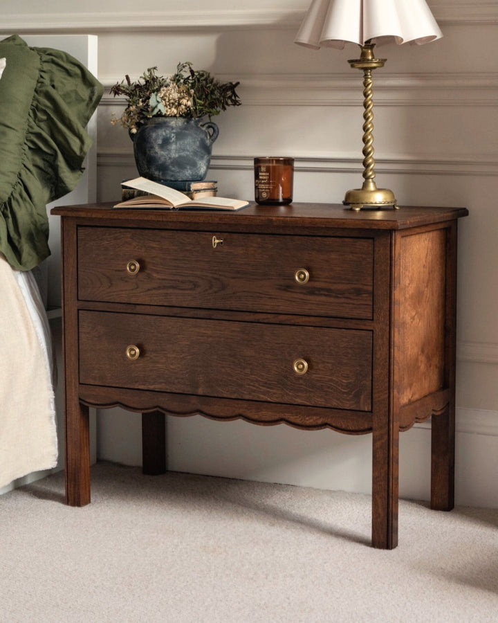 Wooden nightstand with two drawers, lamp, and decorative items in a bedroom setting.