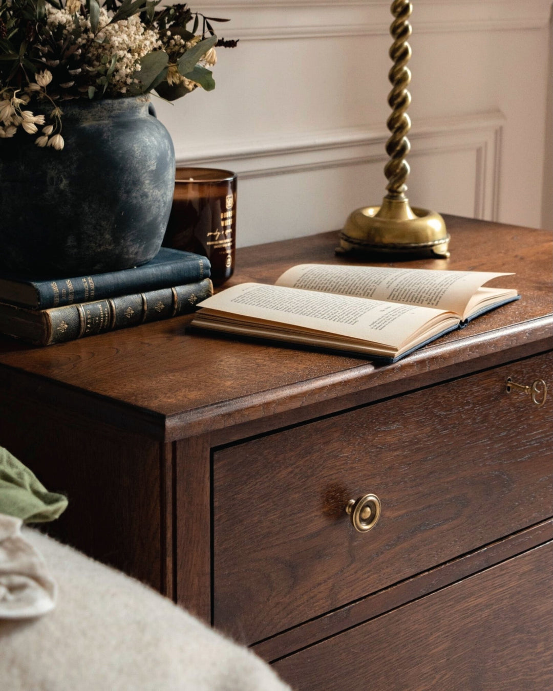 Wooden dresser with open book, vase of flowers, and brass lamp.