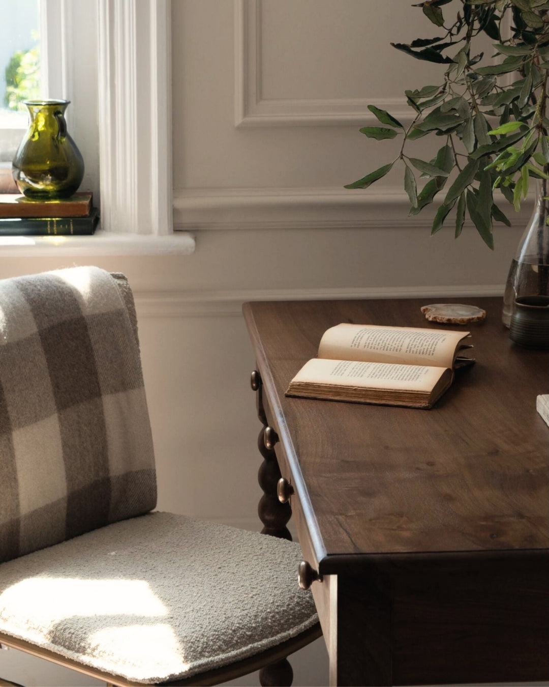 Wooden desk with an open book, chair with plaid cushion, and window with green vase in a home office setting.