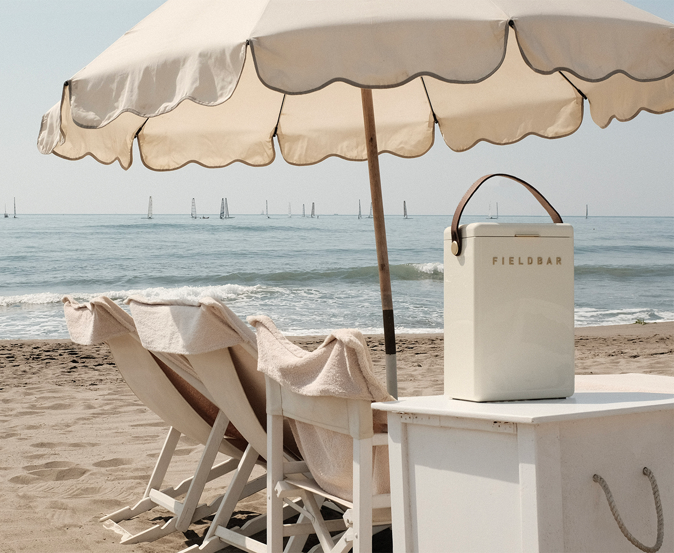 Beach scene with beige umbrella, chairs, and Fieldbar cooler on a sandy shore.
