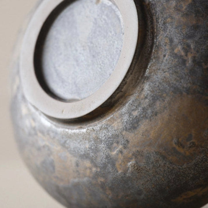 Close up of a handmade ceramic vase with a black and gold color scheme, with a round shape, curved handle and a visible textured surface.