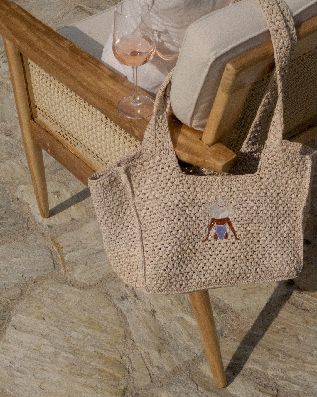 Woven bag with a embroidered design on a stone floor with chairs and a glass in the background.