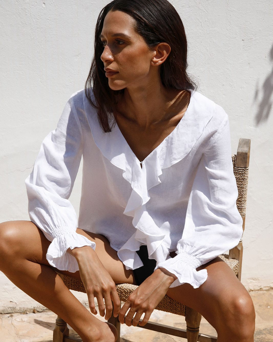 Woman wearing a white blouse sitting on a wooden chair against a light-colored wall.