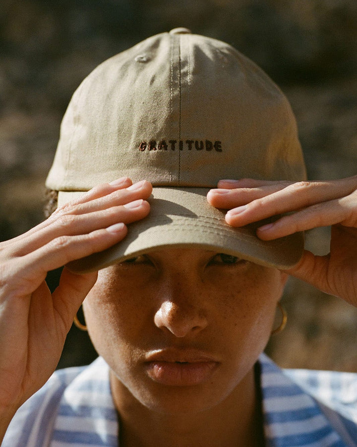 Person wearing a beige cap with 'GRATITUDE' text, holding it on their head.
