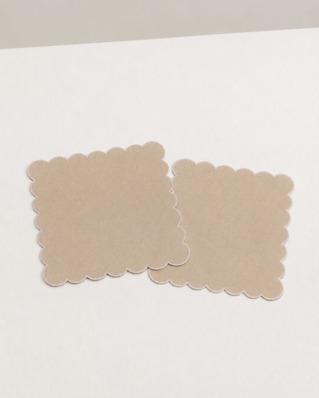 Two beige square napkins  with scalloped edges on a white surface.
