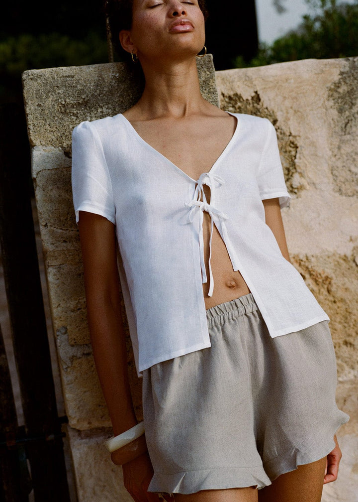 Woman wearing a white top and beige shorts against a stone wall.