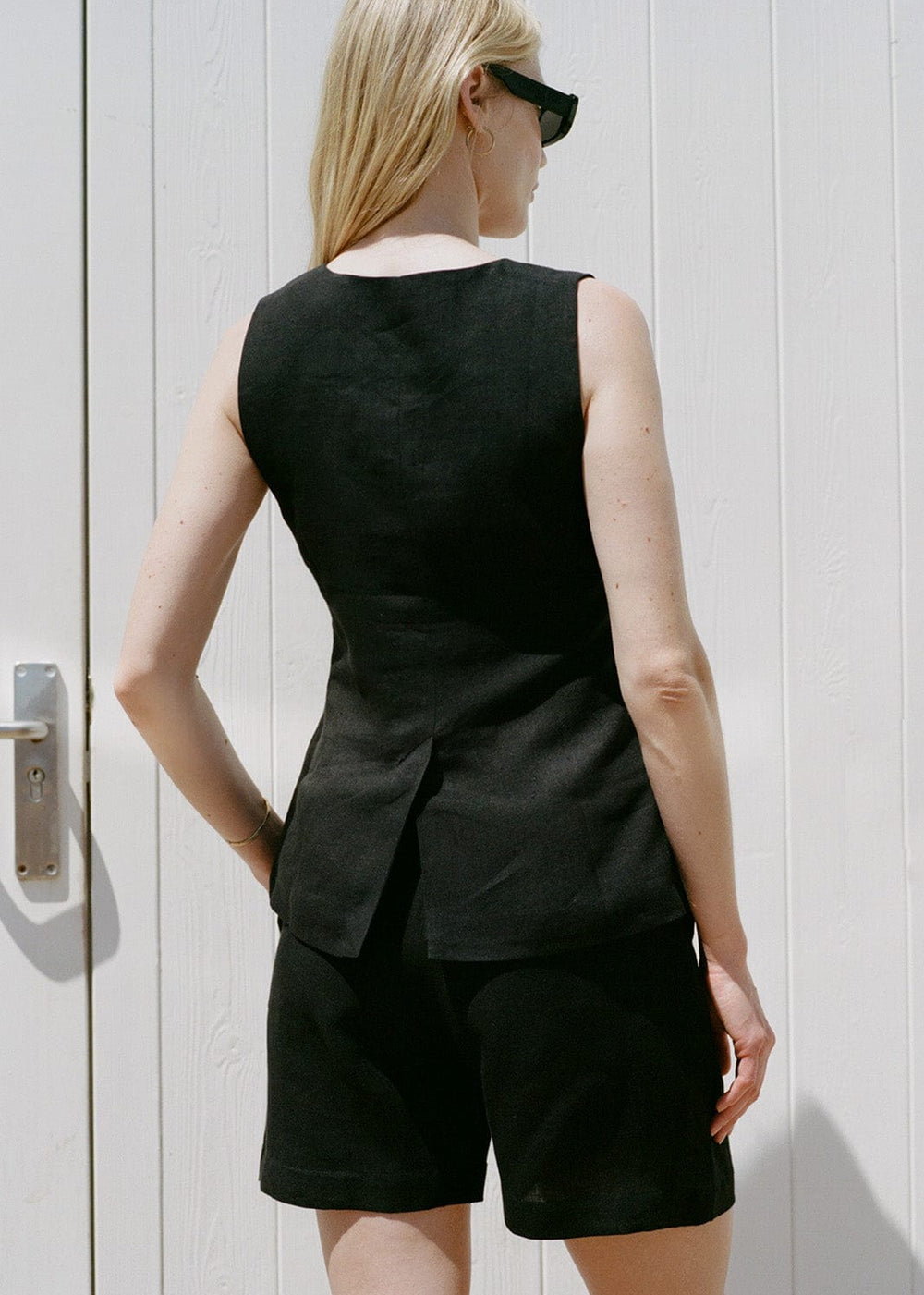 Woman wearing a black sleeveless top and shorts standing against a white wall.
