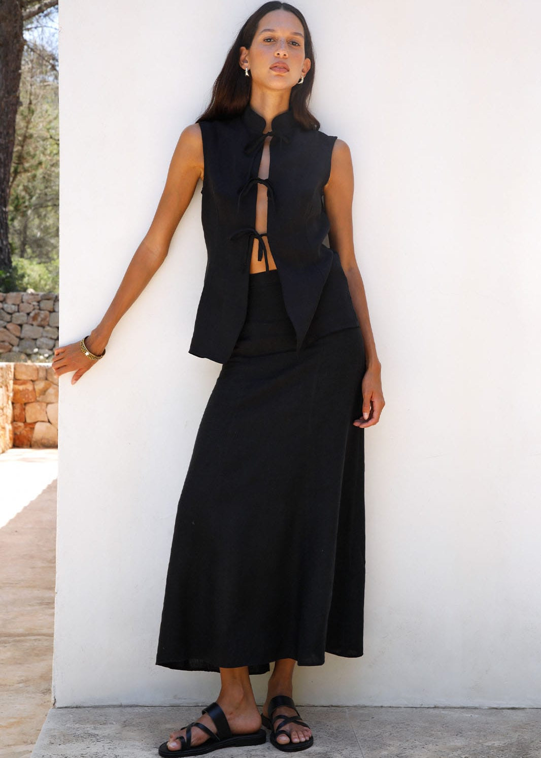 Woman wearing a black sleeveless top and long skirt against a white wall.