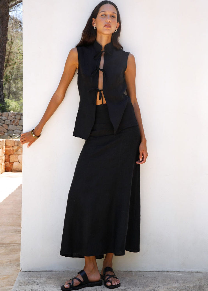 Woman wearing a black sleeveless top and long skirt against a white wall.