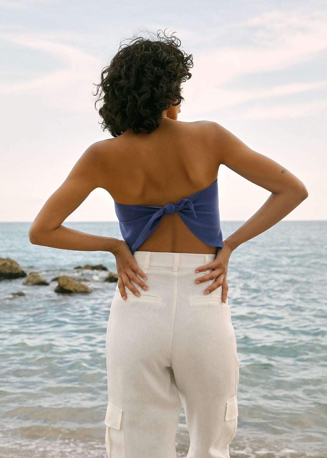 Woman standing on a beach wearing a blue strapless top and white pants, facing the ocean.