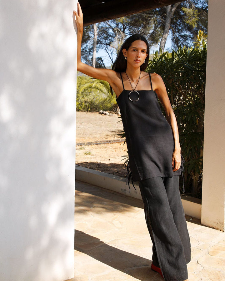 Woman in a black strappy top and black pants standing outdoors near a white wall.