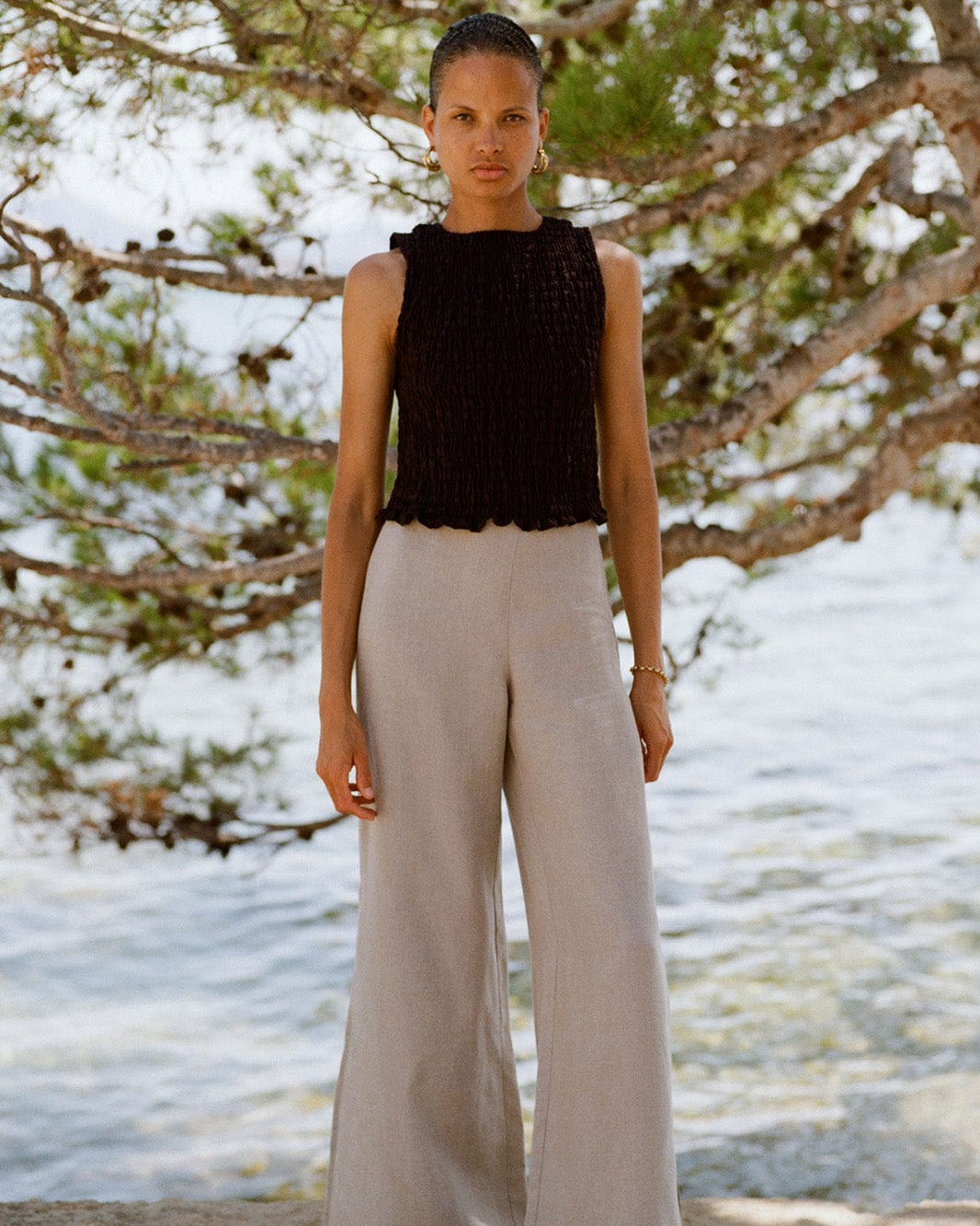 Woman wearing a black sleeveless top and beige pants standing in front of a beach landscape with trees.