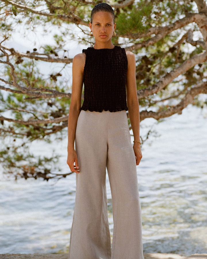 Woman wearing a black sleeveless top and beige pants standing in front of a beach landscape with trees.