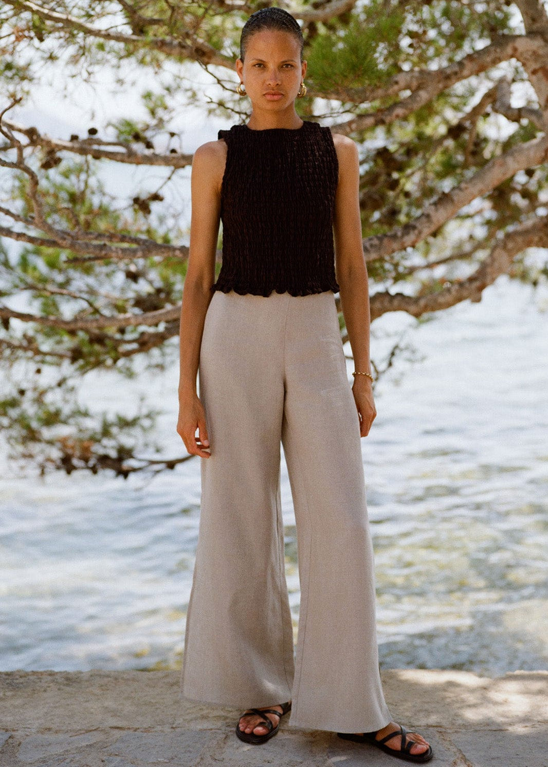 Woman wearing a black sleeveless top and beige wide-leg pants standing in front of a beach landscape.