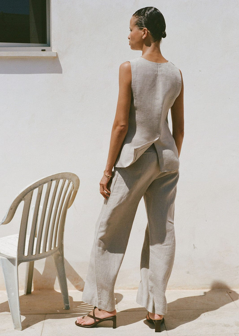 Woman wearing a sleeveless top and beige pants standing against a white wall with a chair nearby.