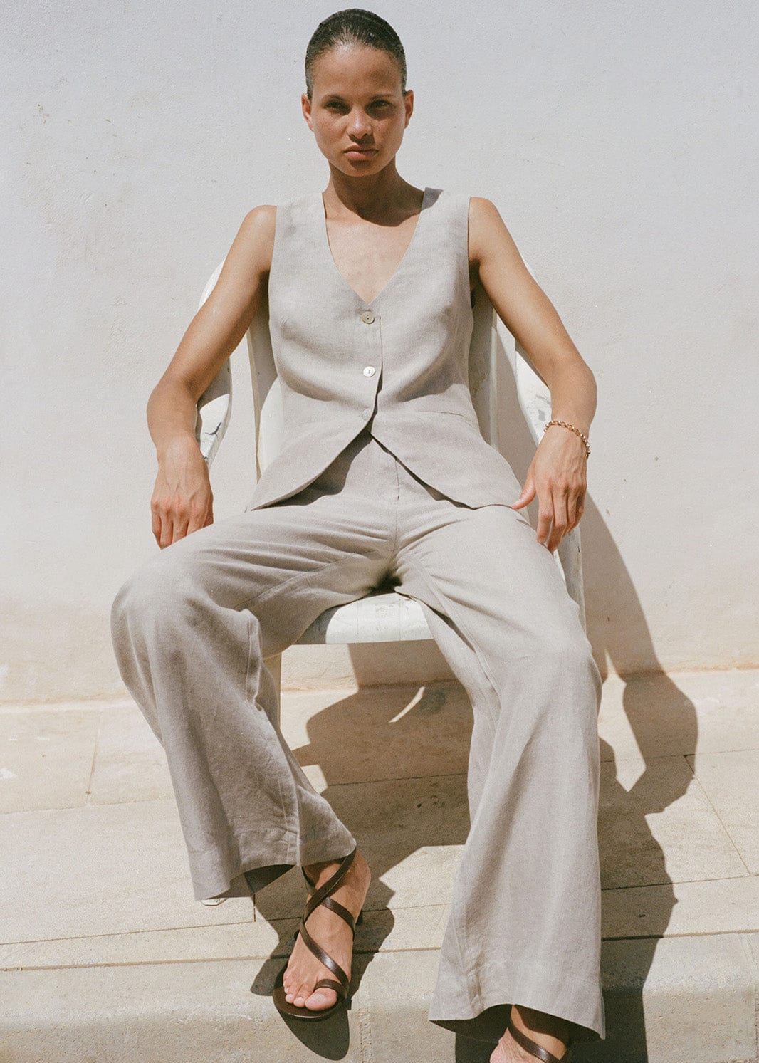 Woman wearing a beige sleeveless top and matching trousers sitting on a stone bench.