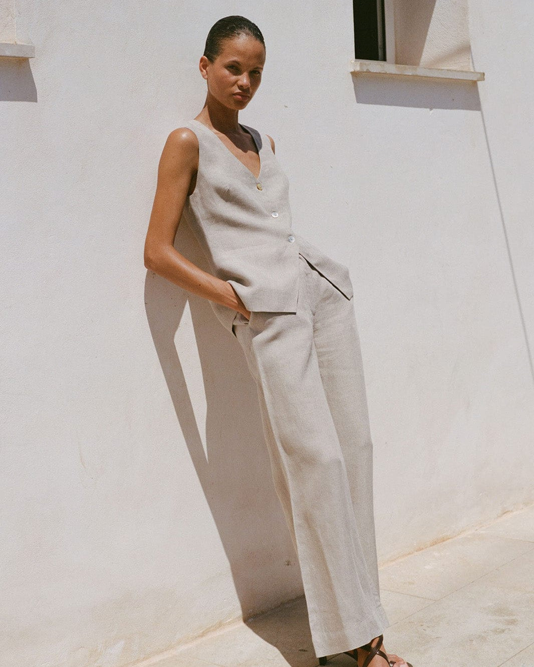 Woman wearing a beige sleeveless top and pants against a white wall.