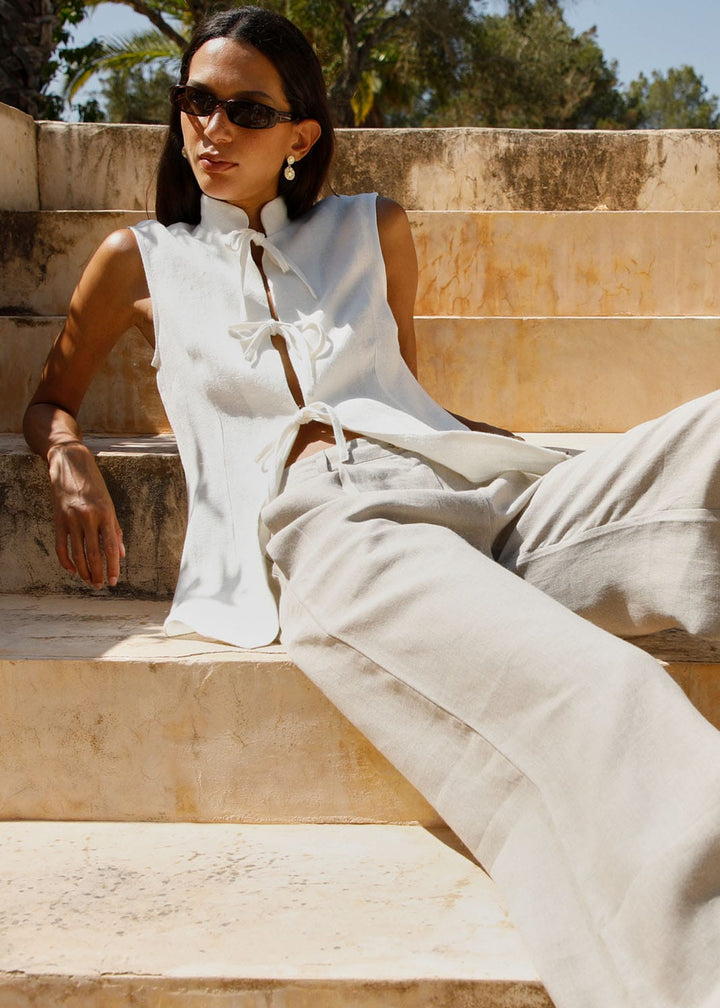 Woman in a white sleeveless top and beige pants sitting on stone steps outdoors.
