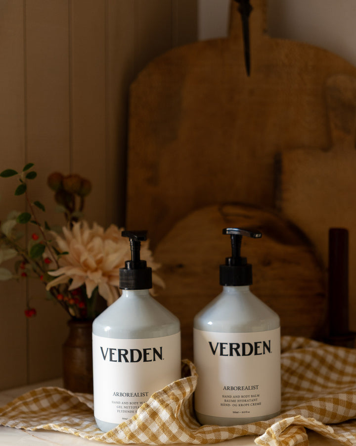 Two large white pump bottles labeled VERDEN ARROWFAIST, placed on a checkered cloth in a styled interior setting with wood tones and flowers in the background.