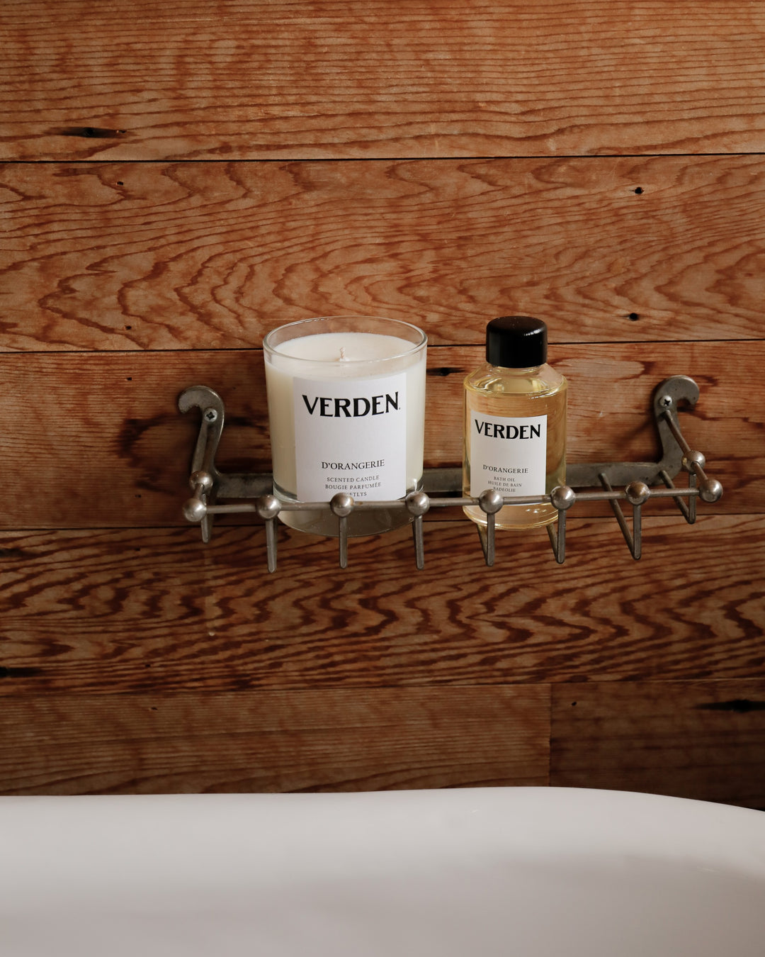Candle and bottle labeled 'VERDEN' on a metal shelf with wooden wall and a bathtub in the foreground.