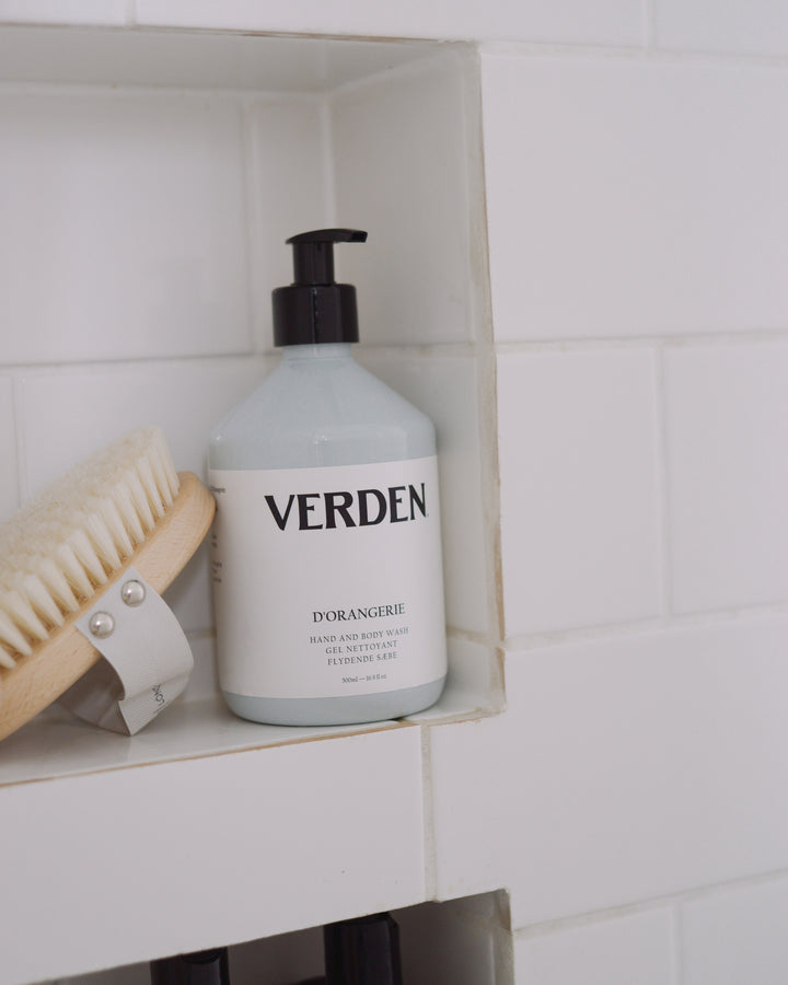 A bottle of VERDEN D'ORANGERIE hand and body wash with a black pump dispenser, resting on a white tiled shelf next to a wooden hair brush.