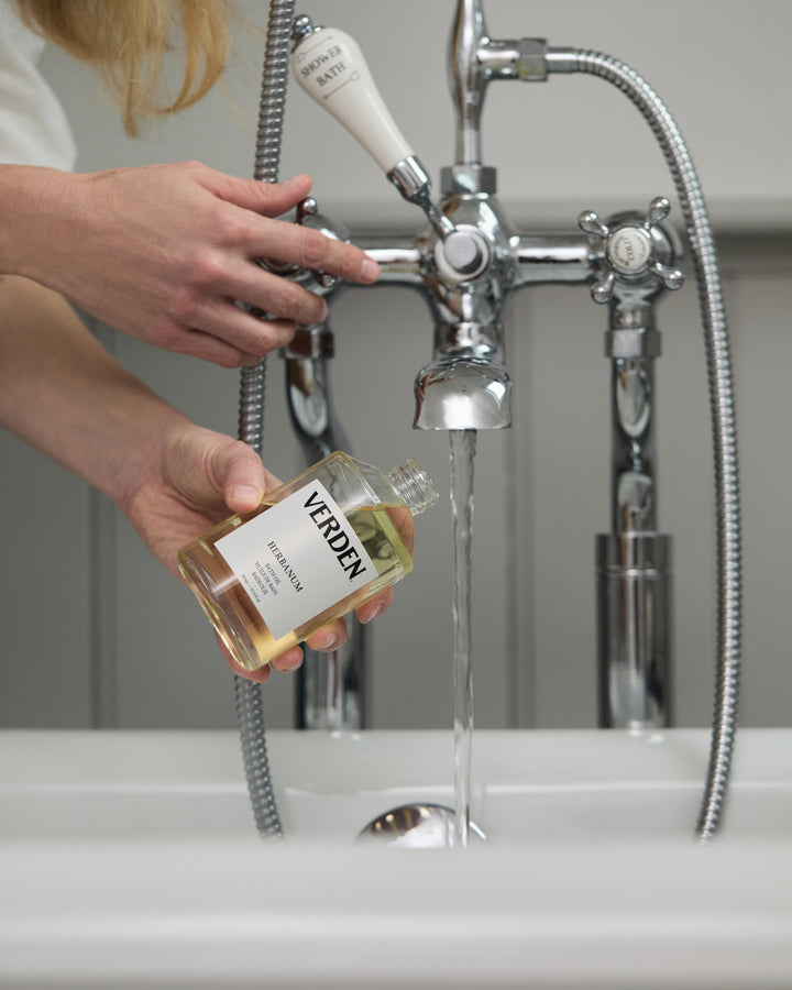 Person pouring liquid from a bottle labeled 'VERDEN' into a sink with a vintage-style faucet.