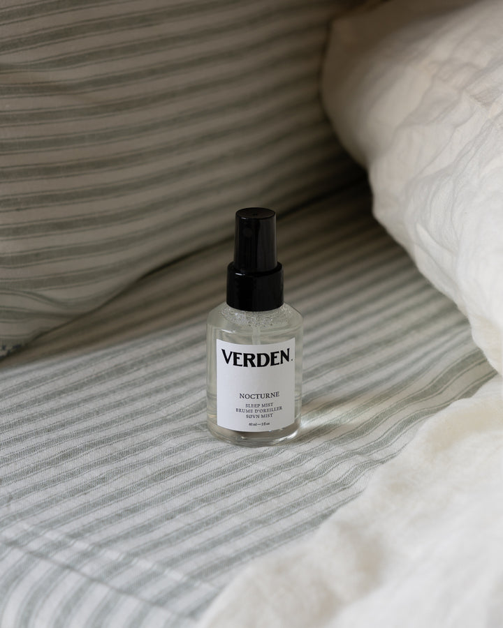 A bottle of VERDEN Nocture body mist with a black spray nozzle and white label, placed on a bed with gray and white striped bedding and white pillows.