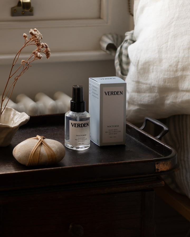 A bottle of VERDEN Nutrient sleep mist and its matching box, styled on a dark wooden tray next to dried flowers and a linen pillow in a bedroom setting.