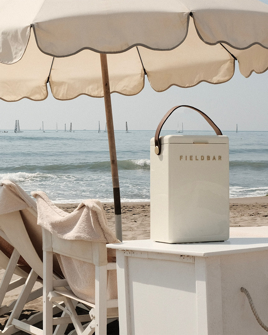 Beige cooler bag with 'FIELDBAR' branding on a table under a large umbrella by the ocean.