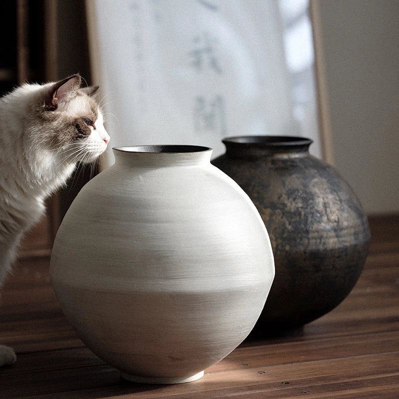 Cat sitting next to a large white vase on a wooden surface with a blurred background